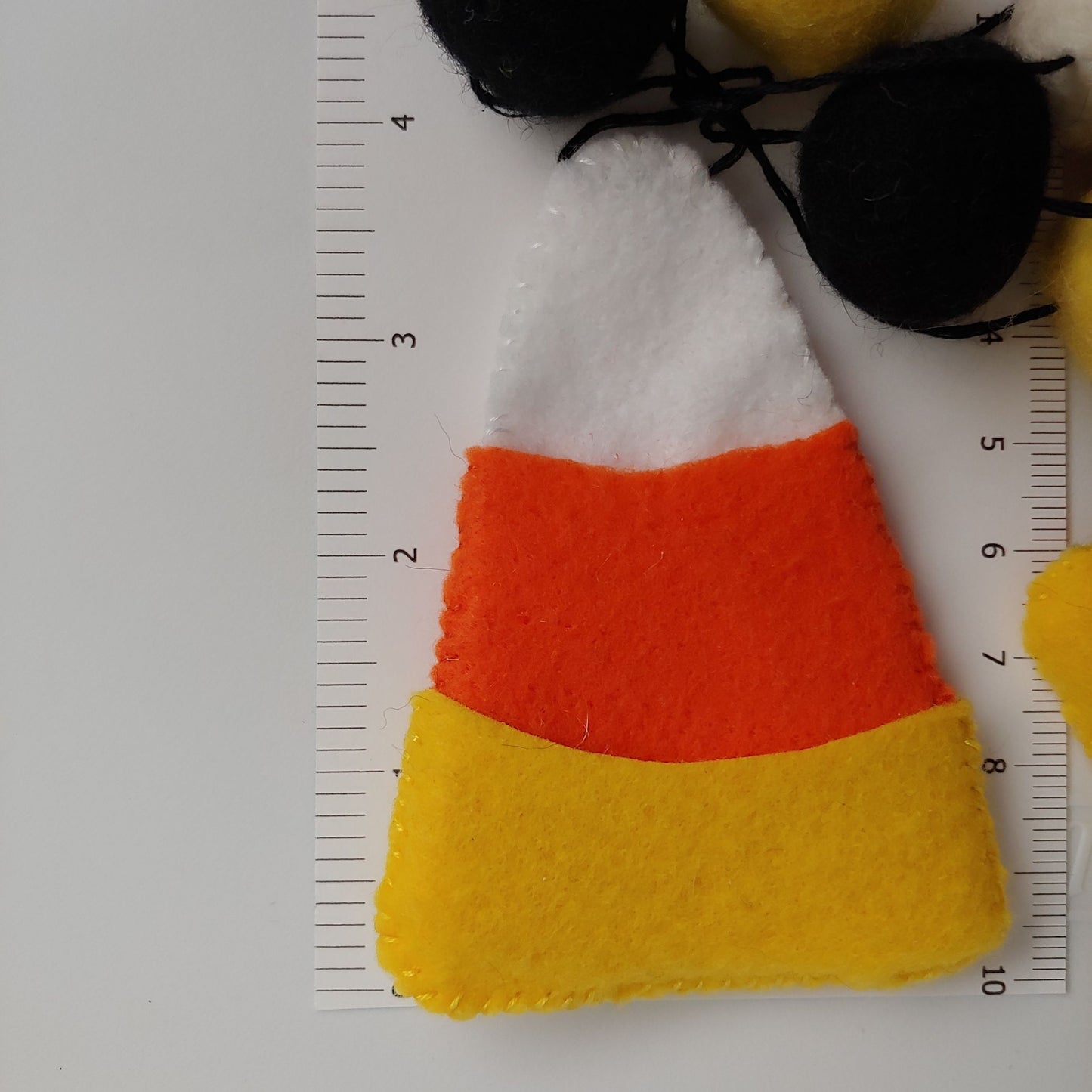 Handcrafted Candy Corn Felt Garland featuring yellow, orange, white, and black felt candy corn pieces with felt balls against ruler. A festive Halloween and fall decoration.