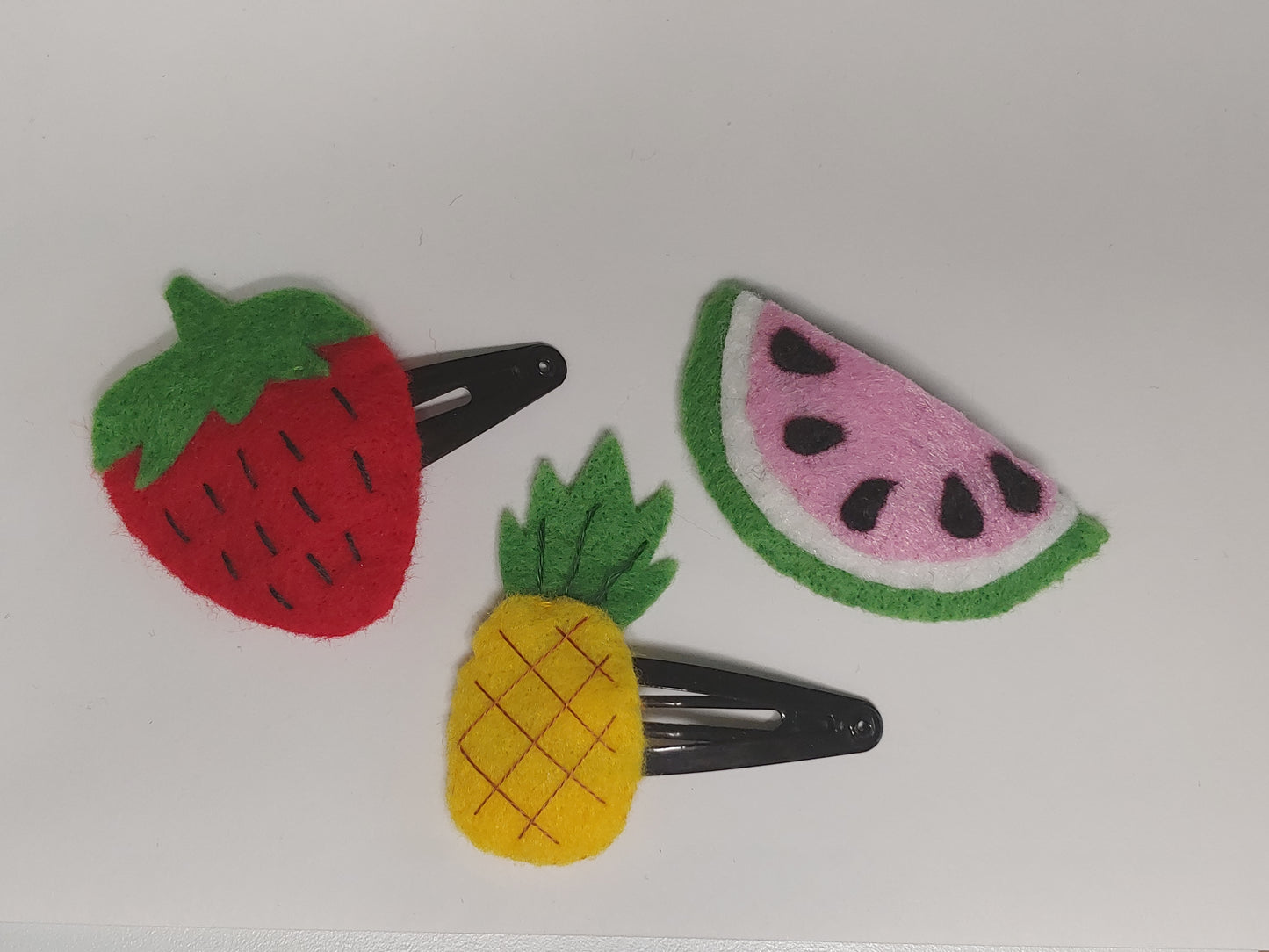 Handmade felt fruit hair clips featuring Watermelon, Pineapple, and Strawberry designs on metal snap clips. A playful, colorful accessory for all ages.