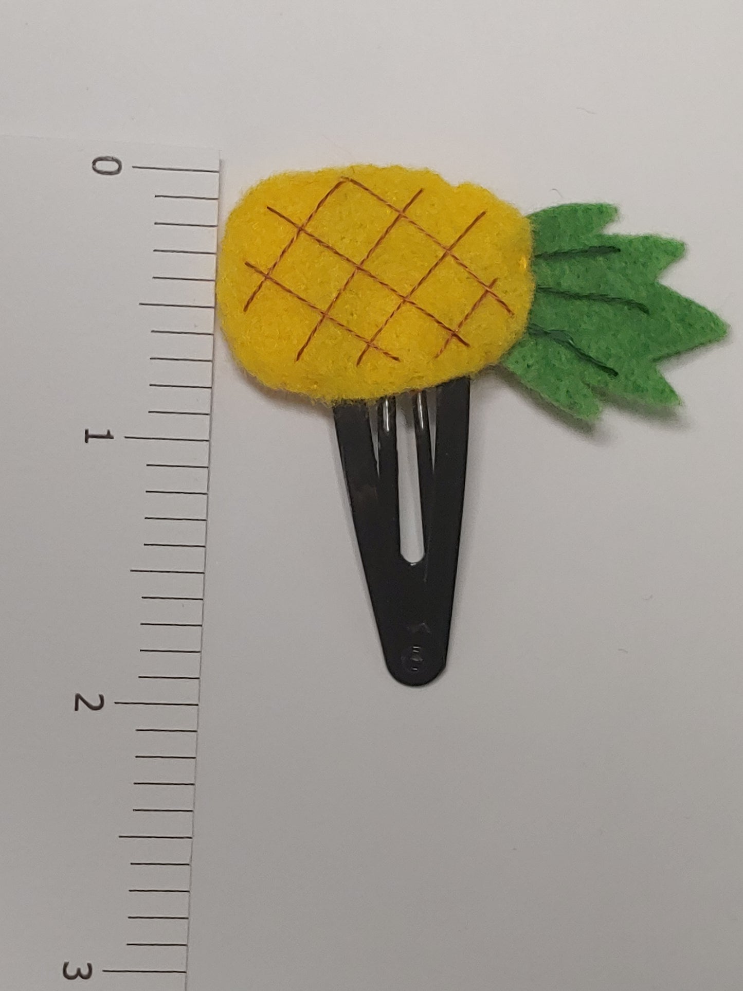Handmade felt fruit hair clip featuring Pineapple design on metal snap clips. Positioned next to ruler.