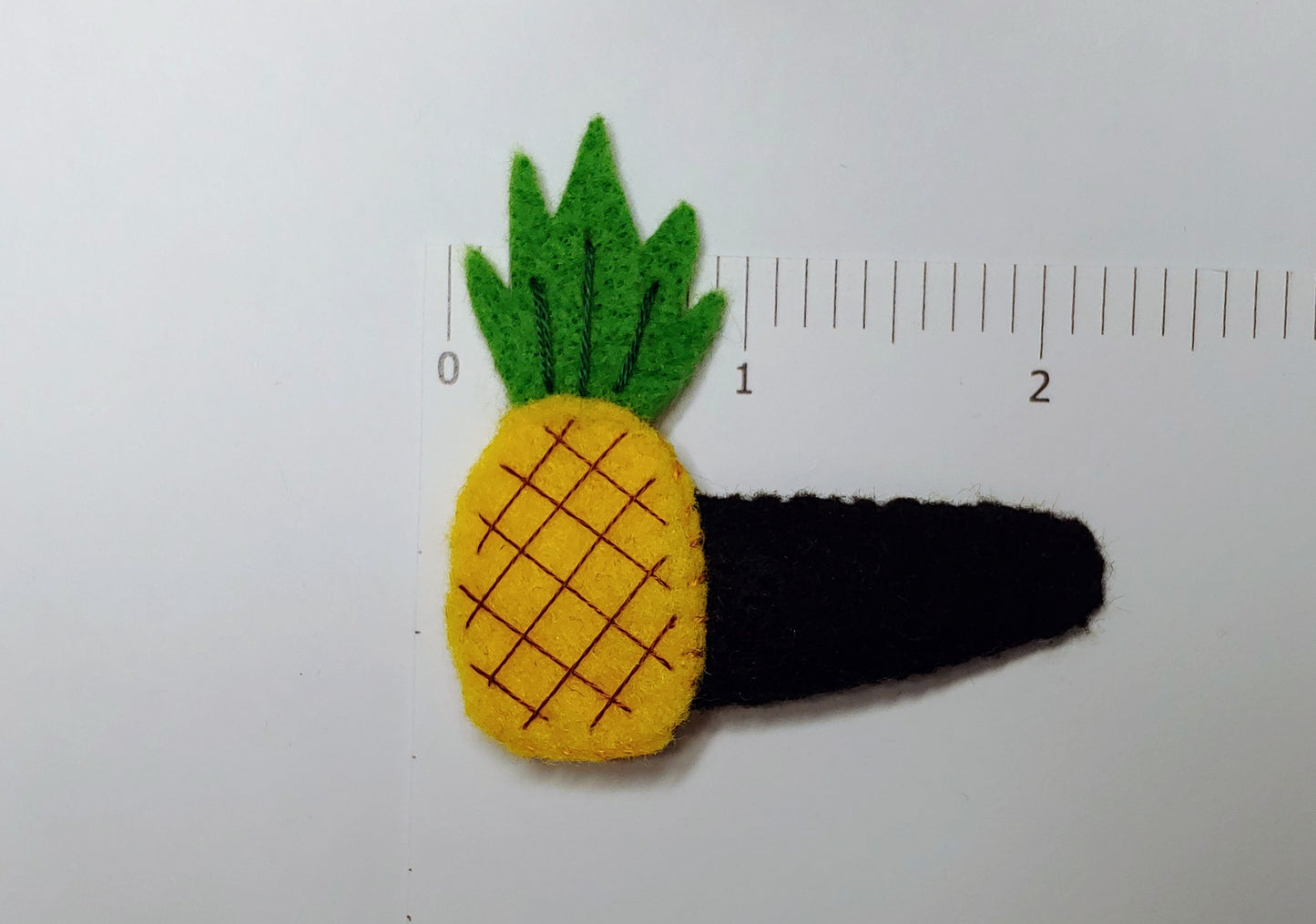 Handmade felt Pineapple Hair Clip with bright yellow and green felt, brown stitched details, and a secure snap clip. Product next to ruler.