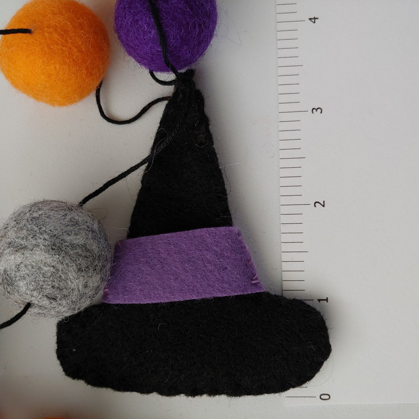 Handcrafted Witches Hat Felt Garland featuring purple, orange, black, and gray felt hats and felt balls - next to ruler. A whimsical Halloween decoration.