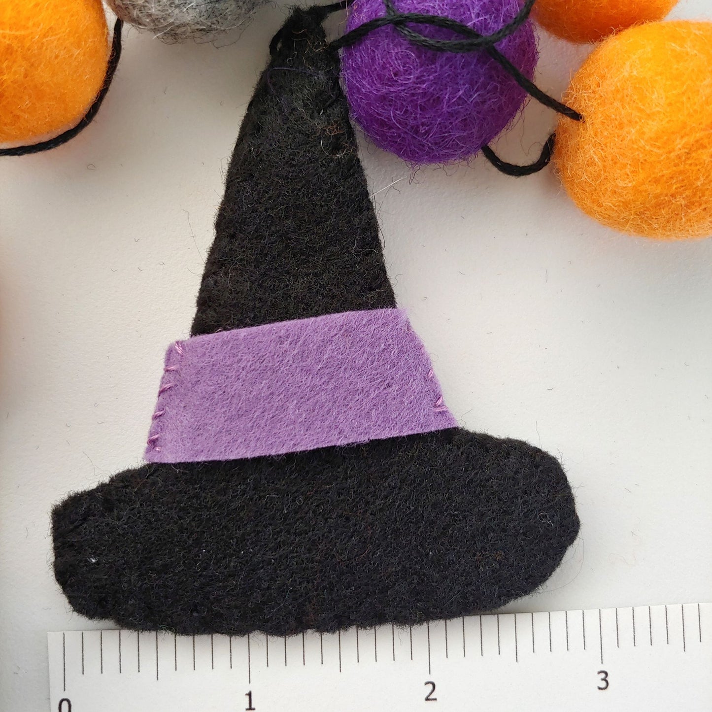 Handcrafted Witches Hat Felt Garland featuring purple, orange, black, and gray felt hats and felt balls - next to ruler. A whimsical Halloween decoration.