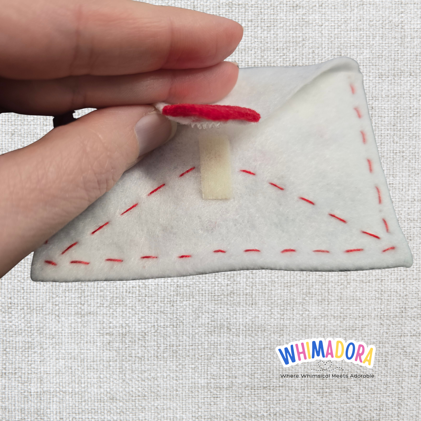 Hand holding a small felt pizza toy with red and brown details on a textured gray background, featuring the Whimadora brand.