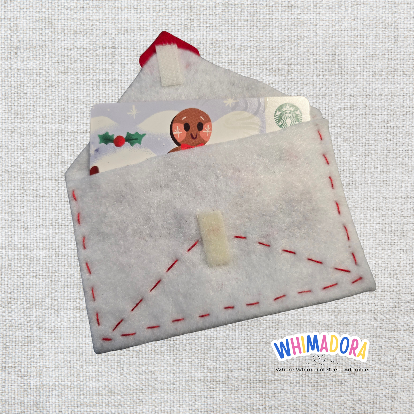 Felt envelope with red stitching and a Starbucks card stuck inside on a textured gray background, branded 'WHIMADORA'.