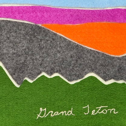 Colorful felt landscape artwork depicting the Grand Teton mountain range. The layered design features green foreground hills, jagged gray mountains, a vibrant orange and pink sunset sky, and a soft blue upper sky. The words “Grand Teton” are stitched in white thread along the bottom.