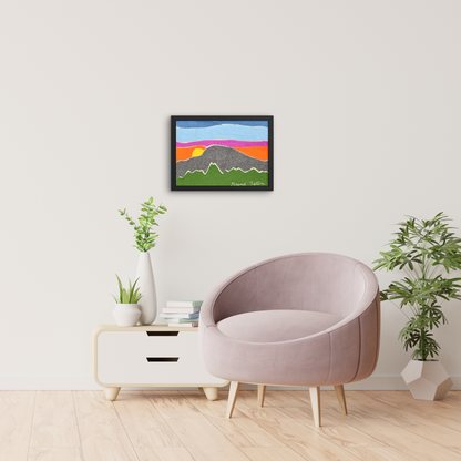 A cozy modern living room with a round blush pink chair, minimalist white side table, and green houseplants. Above the chair, a colorful textile wall art featuring layered mountain silhouettes and a vibrant sunset in hues of orange, pink, purple, and blue is framed in black and displayed on the wall. The artwork has "Grand Teton" stitched in the bottom corner.