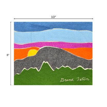 Handmade felt art piece titled ‘Grand Teton,’ measuring 10 inches wide by 8 inches tall. The artwork features layered felt in bold colors depicting a mountain landscape with a yellow sun setting behind dark gray mountains, vivid green foreground, and a multicolored sky in orange, pink, purple, and blue. The words ‘Grand Teton’ are embroidered in white in the bottom right corner.