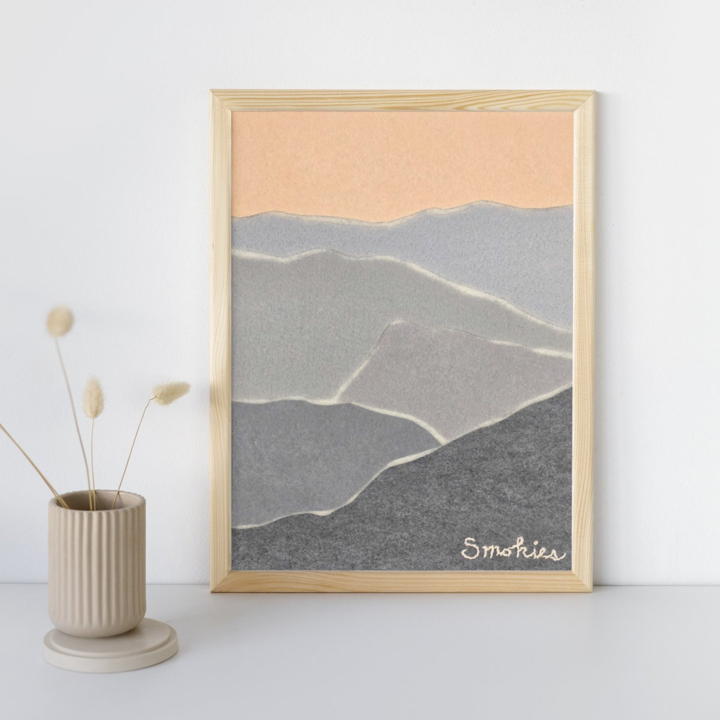 Neutral colors layered to create a "smoky" mountain scene in a neutral frame leaning against a white while with tan vase next to it with 4 stems.