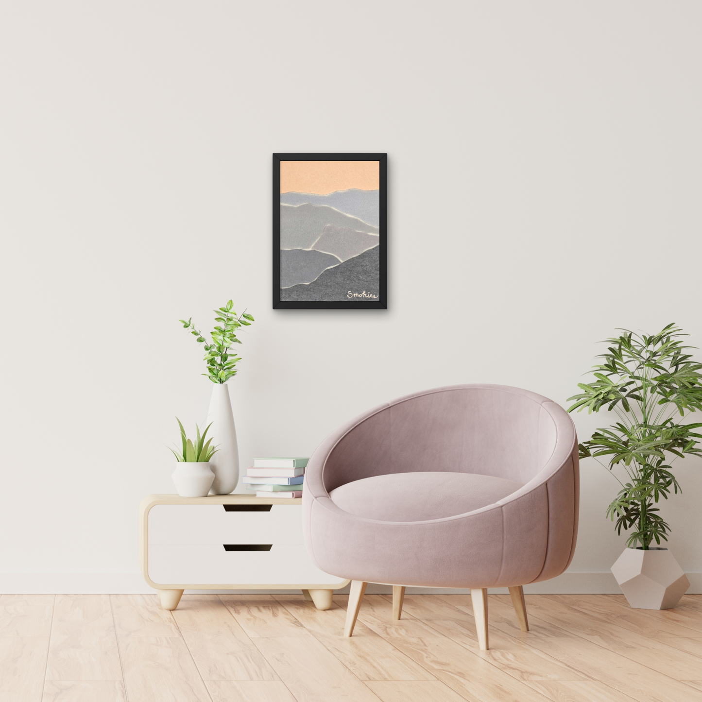 Abstract smoky mountain artwork in black frame on white wall with chair and table with books and plants below it. 