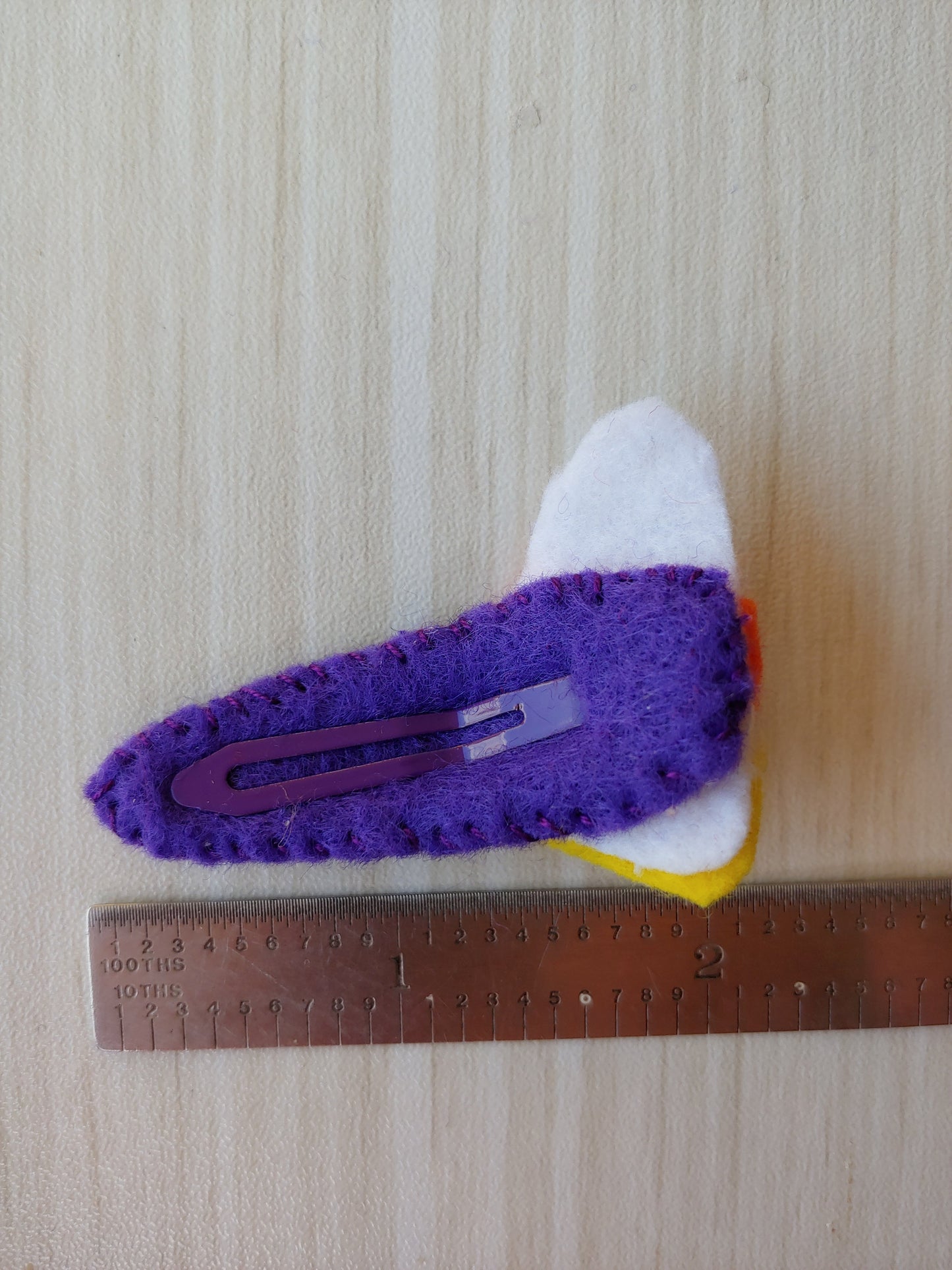 Candy Corn shaped cutout attached to snap clip covered in purple felt. Length of clip 22 tenths.